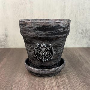 May include: A black plant pot with a distressed silver finish and a matching saucer. The exterior features a centered, raised silver emblem depicting a winged gargoyle-like creature with a crown, set against the dark, textured surface of the pot.