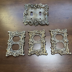 May include: A set of four metallic wall plates with an antique brass finish and ornate floral relief detailing. One plate features two toggle switch openings, while the other three plates each contain two circular electrical outlet openings.