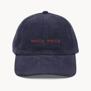May include: A navy blue corduroy baseball cap featuring the text "verily, verily" embroidered in orange thread across the front centre. The cap has a structured crown, a curved brim, and a button at the top, showcasing a textured fabric finish.