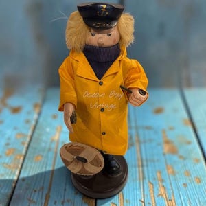 May include: A wooden figurine of a sailor wearing a yellow raincoat, a dark blue turtleneck, and a navy captain's hat with a gold anchor emblem. The figure holds a small pipe and a woven basket, standing on a round base with the text Ocean Boy Vintage.