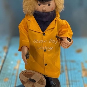 May include: A wooden figurine of a sailor wearing a yellow raincoat, a dark blue turtleneck, and a navy captain's hat with a gold anchor emblem. The figure has blonde hair, holds a black pipe, and stands next to a small tan life preserver.
