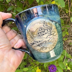 May include: A ceramic mug with a glossy blue and green glaze featuring a circular embossed medallion on the side. The medallion depicts a detailed landscape scene with a cabin, trees, and a small bridge over a stream in shades of tan and brown.
