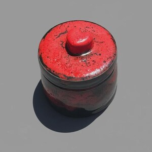 May include: A round ceramic jar with a lid featuring a heart-shaped handle. The exterior displays a textured, mottled glaze in vibrant red and deep black tones, creating a high-contrast finish across the cylindrical body and the circular lid.