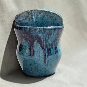 May include: A wall-mounted ceramic planter featuring a glossy glaze with a mottled blend of deep turquoise, light blue, and dark purple streaks. The vessel has a curved, semi-circular top edge and a tapered body with a smooth, reflective finish.