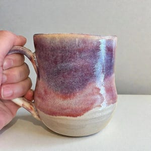 May include: A ceramic mug featuring a glossy glaze with a gradient of deep purple at the top, transitioning into a warm reddish-pink in the center, and ending with a natural cream-colored base. The mug has a curved handle and a smooth finish.