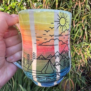 May include: A ceramic mug featuring a hand-painted gradient background transitioning from yellow at the top to orange, red, green, and blue at the base. The mug is decorated with black line art depicting a sun, clouds, birds, mountains, and trees.