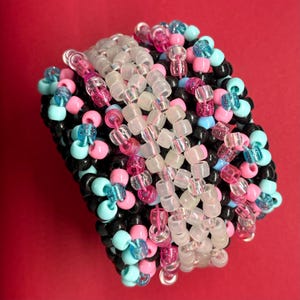 May include: A multi-layered cuff bracelet constructed from numerous plastic pony beads in black, light blue, translucent white, and pink. The beads are arranged in a dense, wide pattern, creating a textured, cylindrical shape with a vibrant color palette.