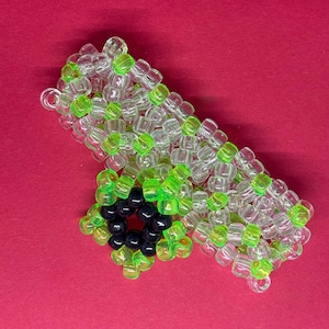 May include: A cuff bracelet constructed from translucent clear and vibrant neon green plastic pony beads. The bracelet features a prominent flower-shaped charm made from neon green beads surrounding a center of six solid black plastic beads.