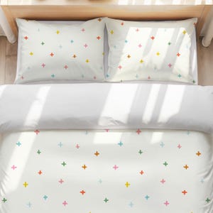 Puede incluir: Un juego de cama blanco que consta de una funda nórdica y dos fundas de almohada, todas con un patrón repetitivo de pequeños signos de más multicolores en tonos rojo, amarillo, verde, azul y rosa dispersos por la superficie de la tela.