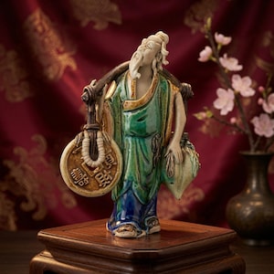May include: A ceramic figurine of a bearded figure wearing a green and blue robe, carrying a wooden yoke across the shoulders. One side of the yoke holds a large circular coin with Chinese characters, and the other side holds a sack-like object.