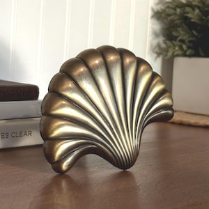 May include: A metallic, brass-toned decorative object shaped like a fan-style seashell. The piece features a series of raised, curved ridges radiating from a central point at the base, with a smooth, polished finish and darkened recessed areas.