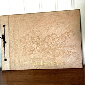May include: A tan rectangular photograph album with a black string binding on the left side. The cover features an embossed illustration of a horse-drawn covered wagon and the word PHOTOGRAPHS printed in gold lettering at the bottom center.