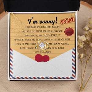 Puede incluir: Un collar dorado con un colgante de nudo y una piedra redonda transparente, presentado en una caja con una tarjeta que dice I'm sorry, un icono de corazón rojo, un sello rojo que dice SORRY y un sello de cera rojo con PLEASE FORGIVE ME.