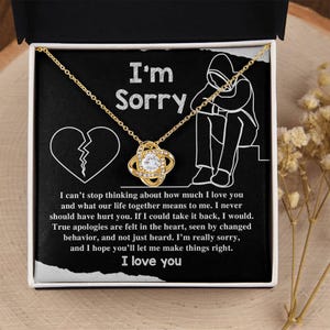Puede incluir: Un collar dorado con un colgante de nudo central con una piedra clara, presentado en una caja negra. El interior muestra el texto I'm Sorry, un gráfico de corazón roto, un dibujo lineal de una persona y un mensaje de disculpa sincero.