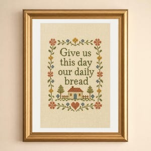 May include: A framed cross-stitch embroidery on beige fabric featuring the text "Give us this day our daily bread" in dark green letters, surrounded by a floral border, a small house, and a red heart, all enclosed in a gold frame.