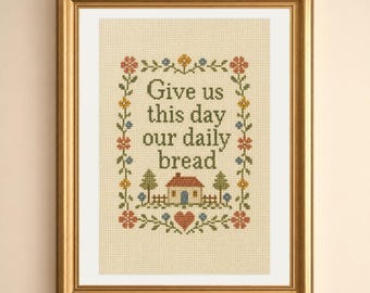 May include: A framed cross-stitch embroidery on beige fabric featuring the text "Give us this day our daily bread" in dark green letters, surrounded by a floral border, a small house, and a red heart, all enclosed in a gold frame.