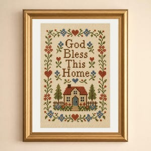 Op de afbeelding: Een ingelijste kruissteek sampler op beige stof met de tekst God Bless This Home in bruine letters. Het ontwerp bevat een centraal huis met een rood dak, groene bomen en een decoratieve rand van rode harten en blauwe en oranje bloemen.