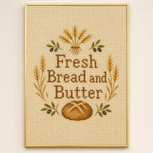 May include: A framed cross-stitch embroidery on beige Aida cloth featuring the text Fresh Bread and Butter in brown thread. The design includes golden wheat stalks, small green olive branches, and a brown loaf of bread at the bottom.