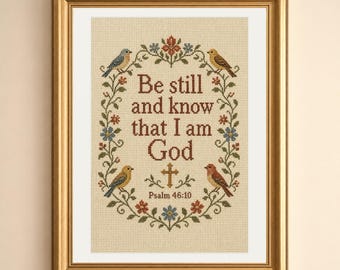 May include: A framed cross-stitch embroidery on beige fabric featuring the text Be still and know that I am God and Psalm 46:10 in brown letters. The design includes a floral wreath with green vines, small red and blue flowers, and four birds.