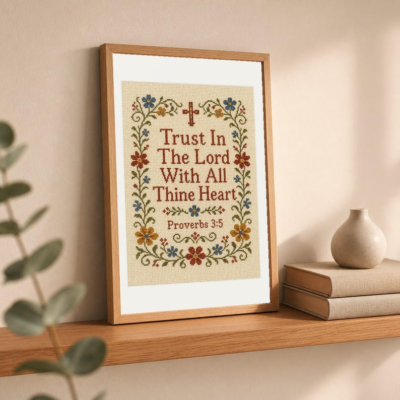 May include: A framed cross-stitch embroidery featuring the text Trust In The Lord With All Thine Heart Proverbs 3:5 in dark red lettering, surrounded by a decorative floral border with blue, red, and yellow flowers and a small cross at the top.
