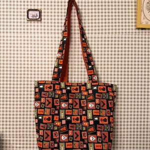 May include: A black fabric tote bag featuring a repeating pattern of orange, green, and white squares. Each square contains Halloween-themed illustrations including skulls, black cats, bats, ghosts, pumpkins, and the text "trick or treat" and "boo".