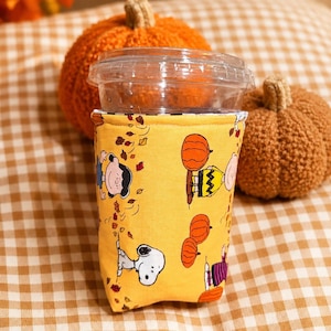May include: A yellow fabric coffee cup sleeve featuring a repeating pattern of Peanuts characters including Snoopy, Charlie Brown, and Lucy, along with orange pumpkins and scattered autumn leaves, wrapped around a clear plastic disposable cup.