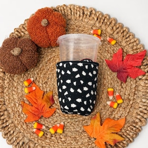May include: A clear plastic cup inside a black fabric sleeve featuring a repeating white ghost pattern. The sleeve is surrounded by two textured orange and brown fabric pumpkins, several orange and red artificial maple leaves, and candy corn.