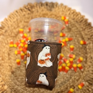 May include: A brown fabric cup sleeve featuring a repeating pattern of white ghosts wearing black hats, each holding an orange book and a jack-o'-lantern candy bucket, wrapped around a clear plastic cup with a visible black circular logo.