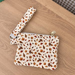May include: A rectangular quilted wristlet pouch featuring a cream-colored fabric printed with a pattern of small brown, orange, and yellow autumn leaves and acorns. The pouch includes a matching fabric wrist strap and a gold-toned metal zipper.