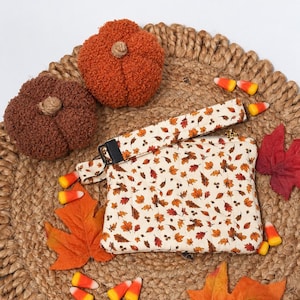 May include: A rectangular fabric pouch with a cream background and a repeating pattern of small autumn leaves and acorns. The pouch features a matching fabric wrist strap attached with a black metal clasp and a zipper closure along the top edge.