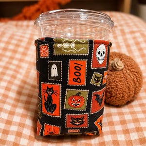 May include: A fabric cup sleeve featuring a black background with a repeating pattern of rectangular postage stamp-style illustrations including a white skeleton, a skull, a ghost, a black cat, a pumpkin, a bat, and the orange text "Boo!".