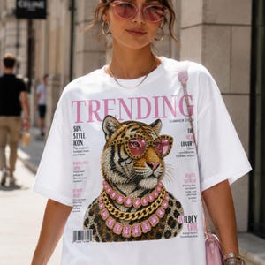 Könnte beinhalten: Ein weißes Kurzarm-T-Shirt mit einem Grafikdruck eines Tigers, der eine rosagetönte Sonnenbrille und geschichtete rosa und goldene Halsketten trägt. Der Druck imitiert ein Magazin-Cover mit dem Text TRENDING, SUMMER 2026 und verschiedenen Textspalten.