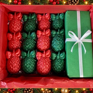 May include: A rectangular gift box containing twelve pinecone-shaped ornaments in alternating red and green colors, each topped with a bow and numbered one through twelve. A separate rectangular green gift box with a white ribbon sits alongside.