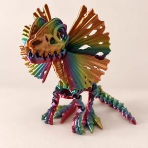 May include: A multi-colored, iridescent dinosaur skeleton figurine featuring a large, flared neck frill. The articulated figure displays a gradient of metallic rainbow hues, including shades of green, blue, purple, and gold, across its entire skeletal structure.