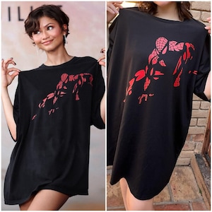 Puede incluir: Una camiseta negra de corte oversize con un estampado gráfico rojo de un personaje inspirado en una araña en posición agachada. La prenda tiene mangas cortas, cuello redondo y un ajuste holgado, con el diseño rojo en la zona del pecho.