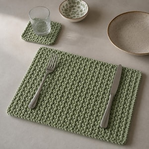 Puede incluir: Un mantel individual rectangular de ganchillo en color verde salvia con un patrón de punto texturizado. Un tenedor y un cuchillo de plata están colocados encima. Un posavasos de ganchillo a juego sostiene un vaso estriado, junto a un cuenco floral.