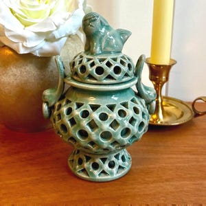 May include: A light green ceramic incense burner featuring an intricate openwork lattice pattern on the body, lid, and base. The lid is topped with a small sculpted creature figurine, and the vessel includes two curved side handles.