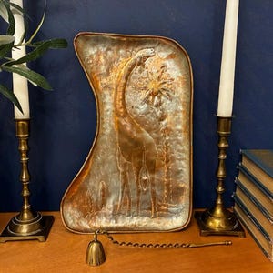 May include: A rectangular copper tray with rounded corners featuring an embossed giraffe and palm tree design. The tray has a metallic finish with varied copper and silver tones. A small brass bell with a twisted handle rests in front of the tray.