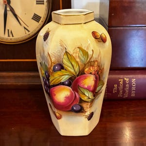 May include: A cream-colored ceramic vase with a hexagonal shape and a gold-trimmed rim. The body features a detailed painted illustration of various fruits, including peaches, grapes, and berries, surrounded by green leaves and small nuts.