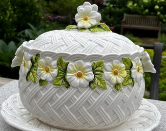 May include: A white ceramic tureen with a lid, featuring a raised basket-weave texture across the surface. The exterior is decorated with three-dimensional white flowers with yellow centers and green leaves, including a flower-shaped handle on the lid.