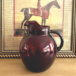 May include: A deep ruby red glass pitcher with a rounded, bulbous body, a wide circular rim, and a curved handle on the side. The pitcher features a prominent pouring spout integrated into the top rim, showcasing a smooth, reflective surface.
