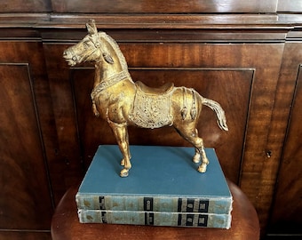 Puede incluir: Una figura de caballo en tono dorado con grabados detallados en la silla y la brida, sobre dos libros azules apilados titulados The Wonder of Knowledge. El caballo presenta una crin y cola texturizadas, con un acabado metálico y adornos.