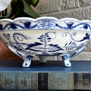 Könnte beinhalten: Eine weiße Porzellanschale mit gewelltem Rand und drei verzierten Füßen, verziert mit einem blauen Blumenmuster aus komplexen Schnörkeln, Blättern und Blüten. Die Schale hat eine glänzende Oberfläche und steht auf einem dunkelblauen Buch.