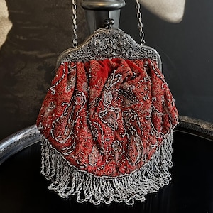 May include: A red velvet handbag featuring intricate silver beadwork in a paisley pattern, a decorative silver metal frame with a kiss-lock clasp, and a long silver chain strap. The bottom edge is adorned with a dense fringe of silver beads.