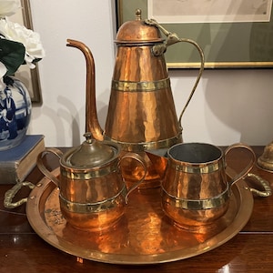 May include: A hammered copper tea set featuring a tall teapot, a lidded sugar bowl, and a creamer, all accented with brass bands. The set sits on a matching round hammered copper tray with brass handles, showcasing a warm metallic finish.