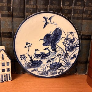 May include: A round ceramic plate featuring a blue and white hand-painted design of two ducks swimming among lotus flowers and lily pads, with a third duck flying above. The plate has a thin blue rim and a glossy finish on the surface.