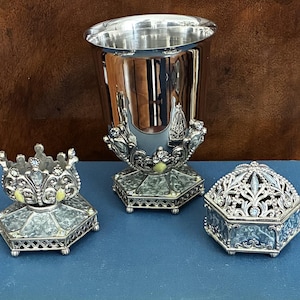 May include: A silver-toned metal set consisting of a tall, polished cup, a small open crown-shaped holder, and a lidded hexagonal box. Each piece features intricate filigree metalwork, light green accents, and small clear embedded crystals.