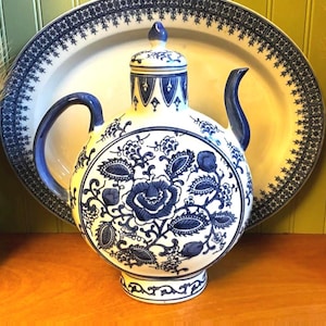 May include: A round ceramic teapot featuring a white base with intricate blue floral patterns, a curved handle, a slender spout, and a matching lid. The teapot has a decorative blue and white border design on the neck and base, showcasing a classic aesthetic.
