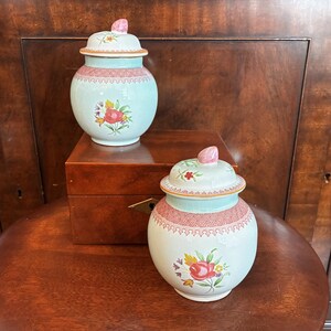 May include: Two matching ceramic ginger jars with lids, featuring a pale mint green base, a red lattice pattern around the neck, and a colourful floral bouquet on the front. Each lid is topped with a small, pink, bud-shaped finial handle.