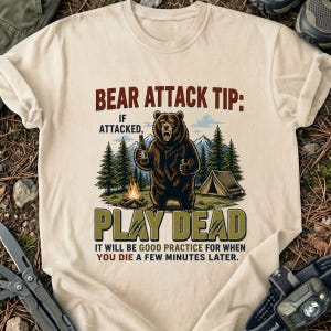 May include: A cream-colored t-shirt featuring a graphic of a brown bear holding a bottle in a forest setting with a tent and campfire. The text reads: BEAR ATTACK TIP: IF ATTACKED, PLAY DEAD. IT WILL BE GOOD PRACTICE FOR WHEN YOU DIE A FEW MINUTES LATER.